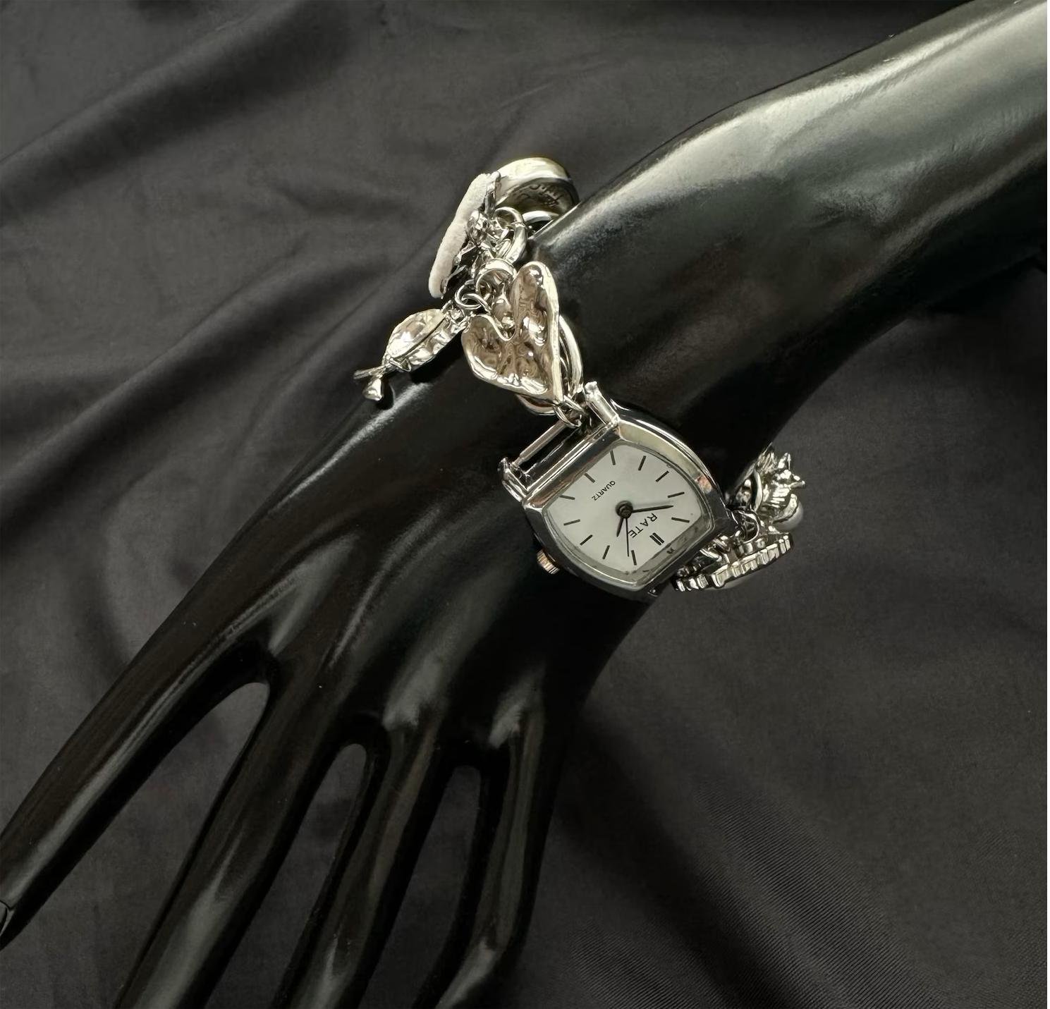 Experimental watch bracelet styled by charm bridges and lateral button mechanisms